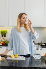 Middle-aged woman squeezes lemon into water and drinks to support vitamins and hydration in home...