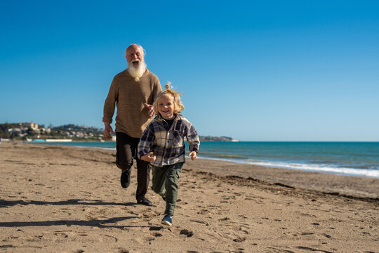 A joyful moment captured as a grandfather and his young grandson run together along the beach. The child, with blonde hair tied up in a small bun, is happily racing beside his grandfather