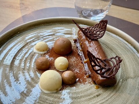 Elegant plated chocolate dessert with glossy ganache bar, cocoa dust, caramel drops and assorted mousse spheres with lace tuile on ceramic plate, fine dining close-up