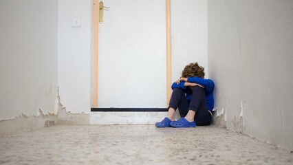 Young boy sitting alone in a corner of a dilapidated room, covering his face, expressing feelings of sadness, loneliness, depression, and neglect, dealing with mental health issues