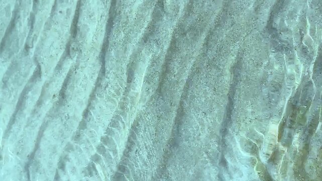 Top-down view of parallel sand ripples on a shallow seabed under clear turquoise water.