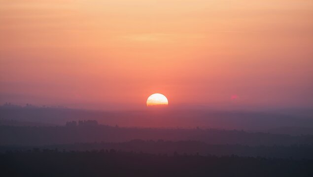 Majestic half sun emerging above layered distant hills creates a breathtaking and colorful orange and pink atmospheric vista during sunrise or sunset.