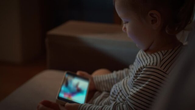 Young child snuggled up on the sofa is completely absorbed in vibrant cartoons on smartphone screen, raising concerns about gadget addiction and potential harm to their eyes and health in the evening