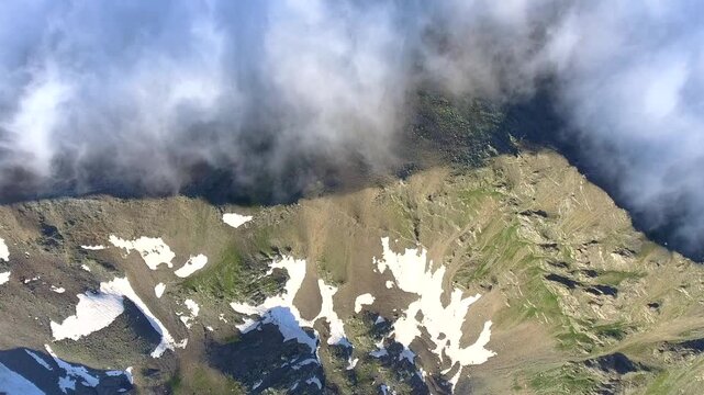 Aerial view of moist air condensing along a high altitude mountain ridge slope in the United States. Orographic cloud formation spreads across rugged alpine terrain under dynamic mountain weather.