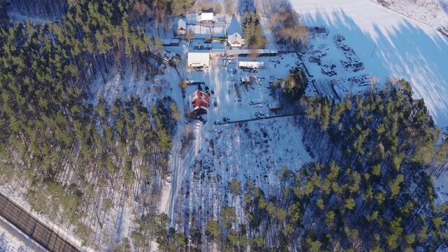 Aerial view of snow covered pines in Poland with a red roof house, outbuildings, farm gear, fields, a red car on a road, tracks in snow, and long winter shadows.