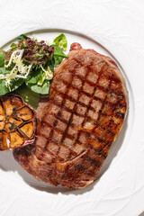 Juicy grilled beef steak served with roasted garlic and fresh green salad on elegant white plate