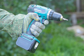 A close-up of a hand holding a cordless drill. The hand is gloved and the drill is turquoise and black. The background is blurred green foliage.