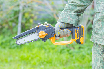 A person holds a battery-powered chainsaw in a green outdoor setting. The tool features a black and orange design, suitable for gardening or landscaping tasks.
