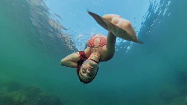 Snorkeling, swimming, underwater. Young woman wearing fins freediving in clear ocean water near coastal cliffs, capturing action selfie style.