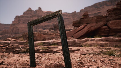 A unique picture frame stands upright in the dry earth, surrounded by red rocks and towering cliffs. The sun shines brightly, highlighting the rugged terrain in a remote desert location. © icetray