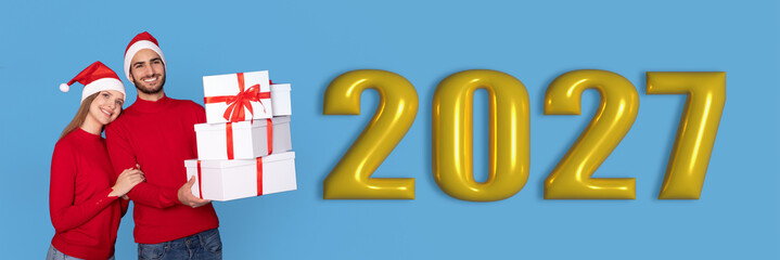 Obraz premium A couple stands together smiling while holding wrapped gifts. They wear matching red sweaters and Santa hats. Behind them is the large number 2027 on a blue background.