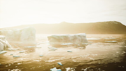 Icebergs drift gently in calm waters, illuminated by the warm glow of early morning light. The landscape features distant hills and a serene atmosphere that enhances the natural beauty. © icetray
