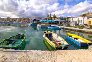 Colourful fishing boats on calm water near church and market stalls at Marsaxlokk harbour, Malta