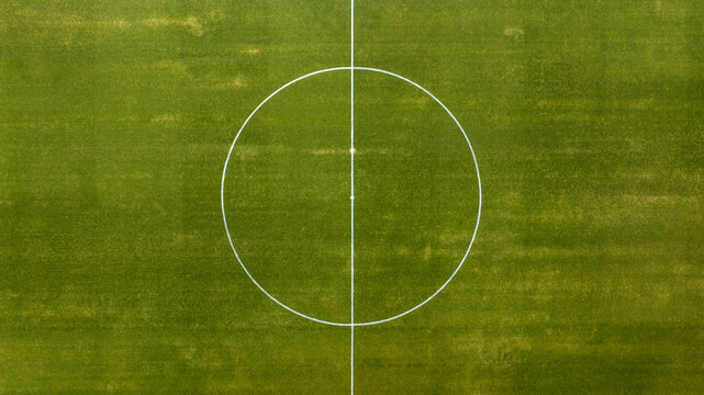 High-angle aerial view of a football pitch's center circle and halfway line, showcasing the precise white markings on a lush, uniform green grass surface. Zenithal shot of a empty soccer ground.