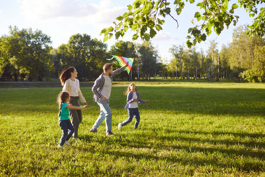 Happy young family with two children running together across green meadow and flying bright kite on sunny day. Smiling parents and kids enjoying active leisure outdoors in nature and having fun.