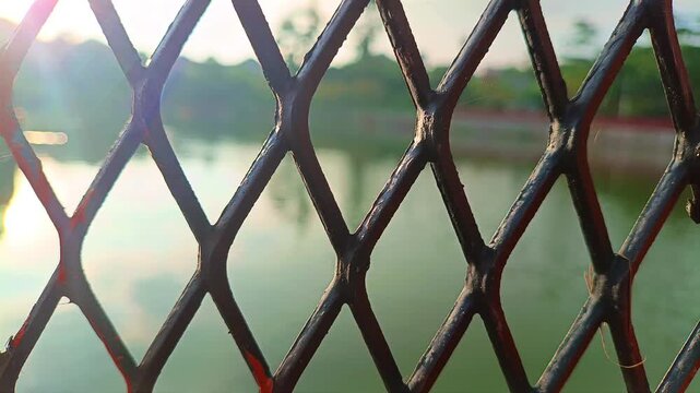 Metal lattice fence pattern with soft sunlight reflection on river water in outdoor environment. Infrastructure concept
