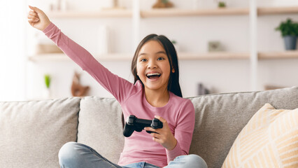 A young girl sits on a couch in a living room, holding a game controller and smiling broadly. She...