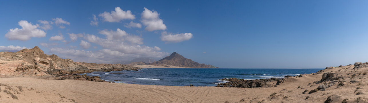 Panoramic beach near Mirbat&ndash;Hasik Road, Oman, with sandy shore, rocky coastline, gentle waves of the Arabian Sea, and distant mountains under a bright blue sky with scattered clouds.