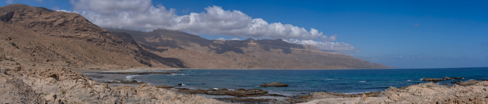Wide coastal panorama along Mirbat&ndash;Hasik Road, Oman: rugged limestone cliffs meeting turquoise Arabian Sea under a clear blue sky with scattered clouds.