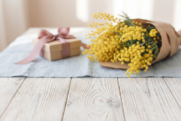 Spring gift box with pink ribbon and yellow mimosa bouquet in paper on wooden table and linen napkin for international Women Day celebration.