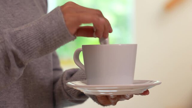 Close-up of hand holding coffee cup and stirring with spoon in bright room, warm sunlight morning atmosphere, relaxing lifestyle and beverage preparation at home.