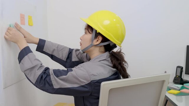 Asian female construction worker is writing or sticking notes onto a corrugated plastic sheet in an office.