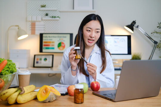 Professional nutritionist explaining vitamin supplements during an online consultation, sitting at a desk with fresh fruits and health charts
