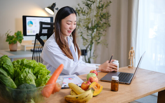 Nutritionist preparing vitamin supplements during an online consultation at a modern home office, with fresh fruits and vegetables on the desk.