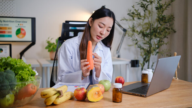 Nutritionist examining a fresh carrot during an online health consultation, surrounded by fruits, vegetables, and dietary supplements in a modern office.