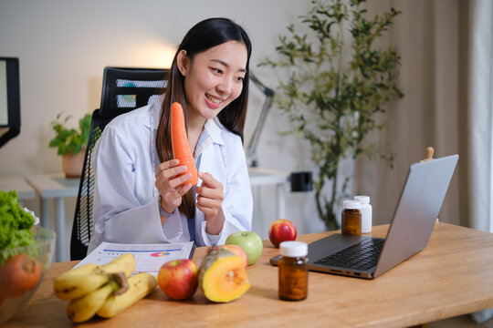 Nutritionist examining a fresh carrot during an online health consultation, surrounded by fruits, vegetables, and dietary supplements in a modern office.