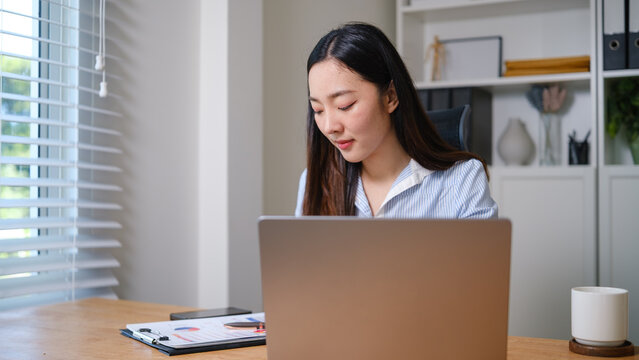Professional businesswoman reviewing financial reports while working on a laptop at her desk in a bright modern office.