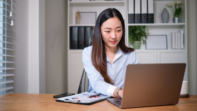Professional businesswoman reviewing financial reports while working on a laptop at her desk in a bright modern office.