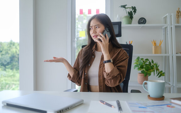 Young businesswoman talking on a smartphone with a confused expression while sitting at her desk in a bright modern office.