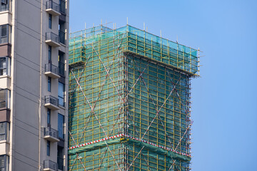 A high-rise building under construction in China, wrapped in scaffolding and green safety netting beside an existing residential tower. High-density housing growth and construction activity © Doublelee