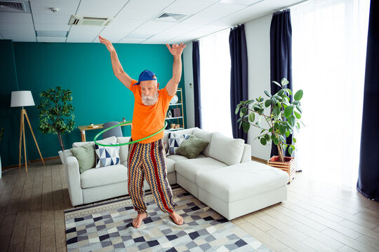 Playful grandpa hula hoops indoors in a cozy living room wearing bright orange shirt and striped pants for a fun exercise moment