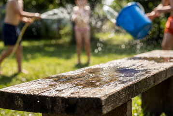 Wet wooden bench in a sunny garden with blurred children splashing water during Polish Lany Poniedziałek tradition.