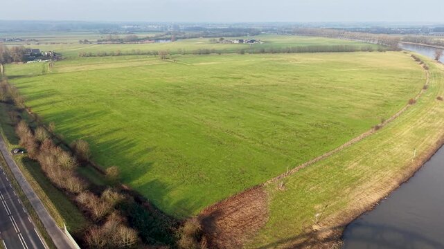 A wide green field stretches between a curving river and a straight roadside, with scattered trees and distant buildings forming a calm, open rural landscape.