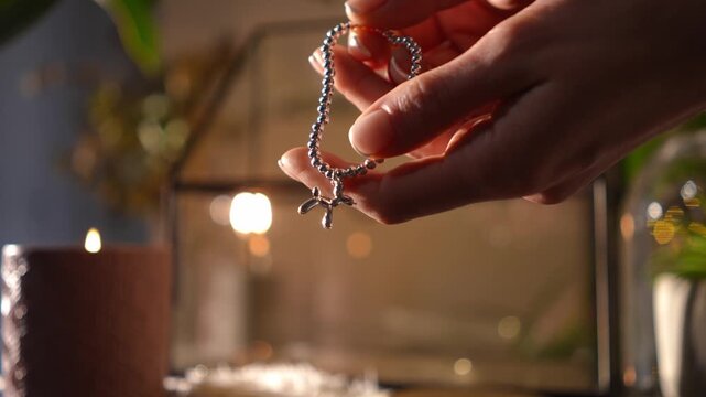 Close-up of female hands holding and inspecting a delicate silver beaded bracelet with a balloon dog charm. The background is warm and cozy