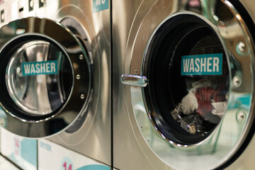 Obraz premium In a public laundry, several washing machines are working. Each machine has clothes inside them. People are nearby, waiting for their laundry to finish. It is daytime