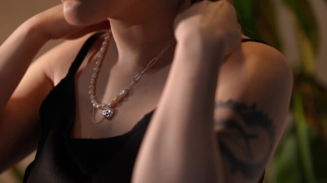 Close-up of a stylish young adult woman putting on an elegant pearl and silver chain necklace with a heart pendant in a softly lit room