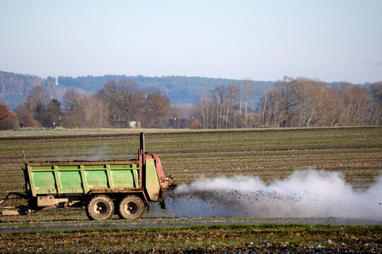 Modern blue tractor pulling a manure spreader trailer, fertilizing a farm field under a clear blue sky. Agricultural machinery at work in the countryside during autumn or spring season.