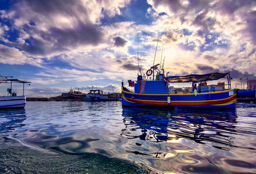 Marsaxlokk, Malta &ndash; Scenic harbor scene with fishing boat, water reflections and atmospheric sky lighting