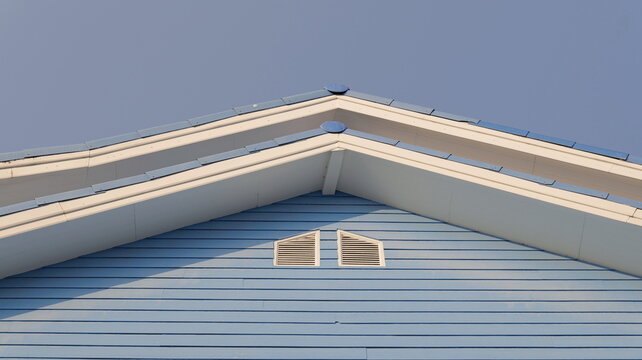A low-angle architectural shot showcases the clean lines of a blue wooden house gable featuring white trim and symmetrical ventilation slats under a clear sky