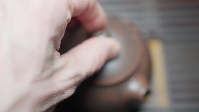Overhead slow motion close-up of a hand placing the lid on a brown clay teapot filled with hot water, completing a step in the traditional tea brewing ritual.