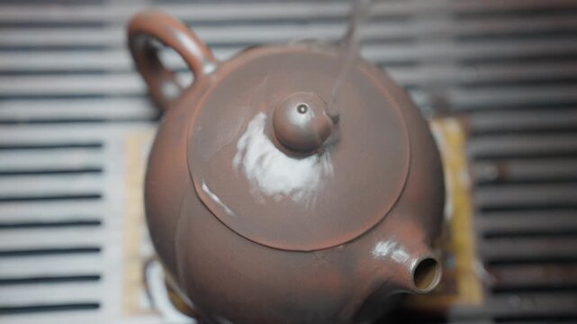 Top-down slow motion close-up. A stream of boiling water is poured over a brown clay teapot on a tea board, generating rising steam and vapor in a tranquil ritual.