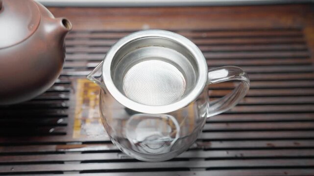 Top-down slow motion of the tea serving ritual. Tea is poured from a brown clay teapot through a strainer into a cha hai (fairness cup) on a bamboo tea board.