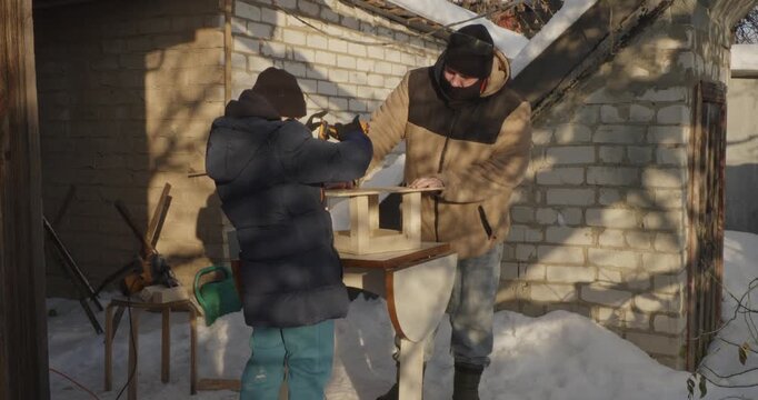 Young son learning from his father how to use a power drill while building a wooden bird feeder together in the backyard during a sunny winter day, fostering family bonding and practical skills.