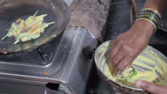 A home chef prepares a spicy Indian snack. The vibrant green chillies are coated in a golden besan batter before being carefully placed onto a black tawa to sizzle and fry into crispy fritters.