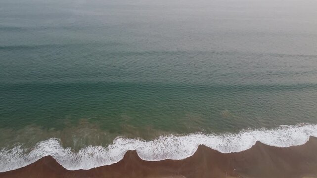 Top-down aerial drone view of gentle ocean waves reaching a sandy beach along the Bay of Bengal coast in Odisha, India. Calm sea water with natural wave patterns and shoreline texture, captured from a