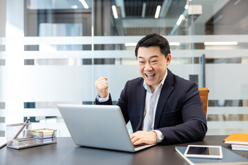 Fototapeta premium Asian businessman, full of joy and enthusiasm, raising his fist in a gesture of triumph while looking at a laptop computer, celebrating achievement and happy news at his modern workspace